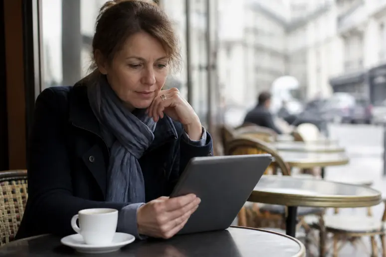 Une femme d'âge mûr sirote un café dans un café parisien, réfléchissant à ses investissements boursiers sur sa tablette.