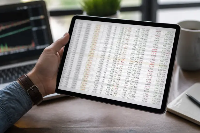 Gros plan sur une tablette affichant des calculs de plus-value pour des actifs numériques, avec une main tenant l'appareil.