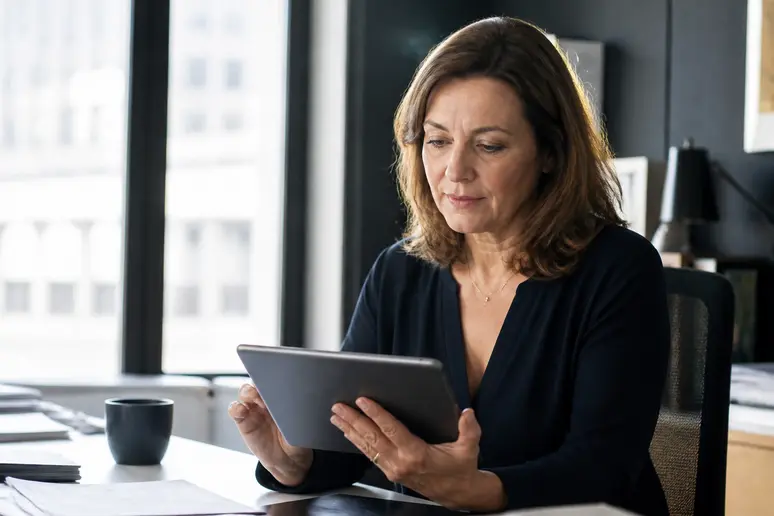 Une femme française mûre dans un bureau, examinant des documents d'investissement sur une tablette avec une lumière naturelle.