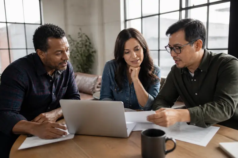 Trois amis échangent des vues sur des documents financiers et un ordinateur portable, reflétant les avantages et inconvénients de différentes approches d'investissement.