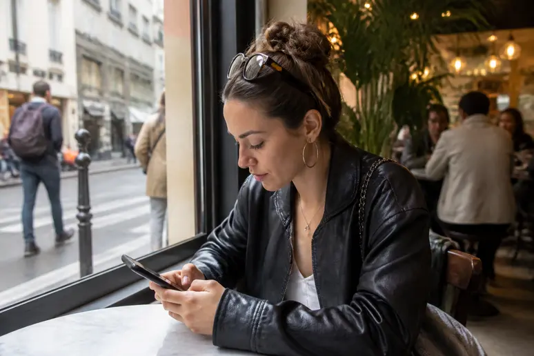 Une jeune femme concentrée sur son smartphone dans un café parisien, analysant les cours des cryptomonnaies du métaverse.