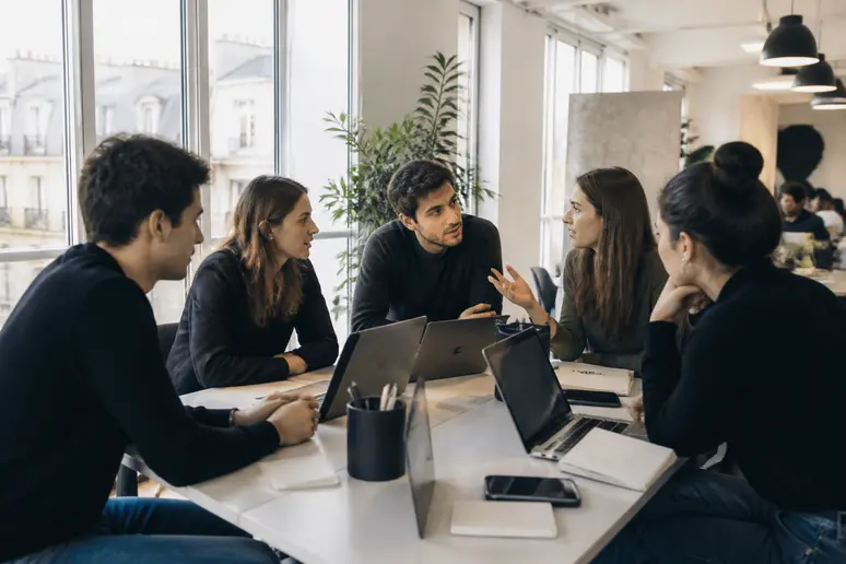 Des jeunes adultes français échangent autour d'une table dans un espace de coworking, discutant de concepts numériques sur leurs écrans.